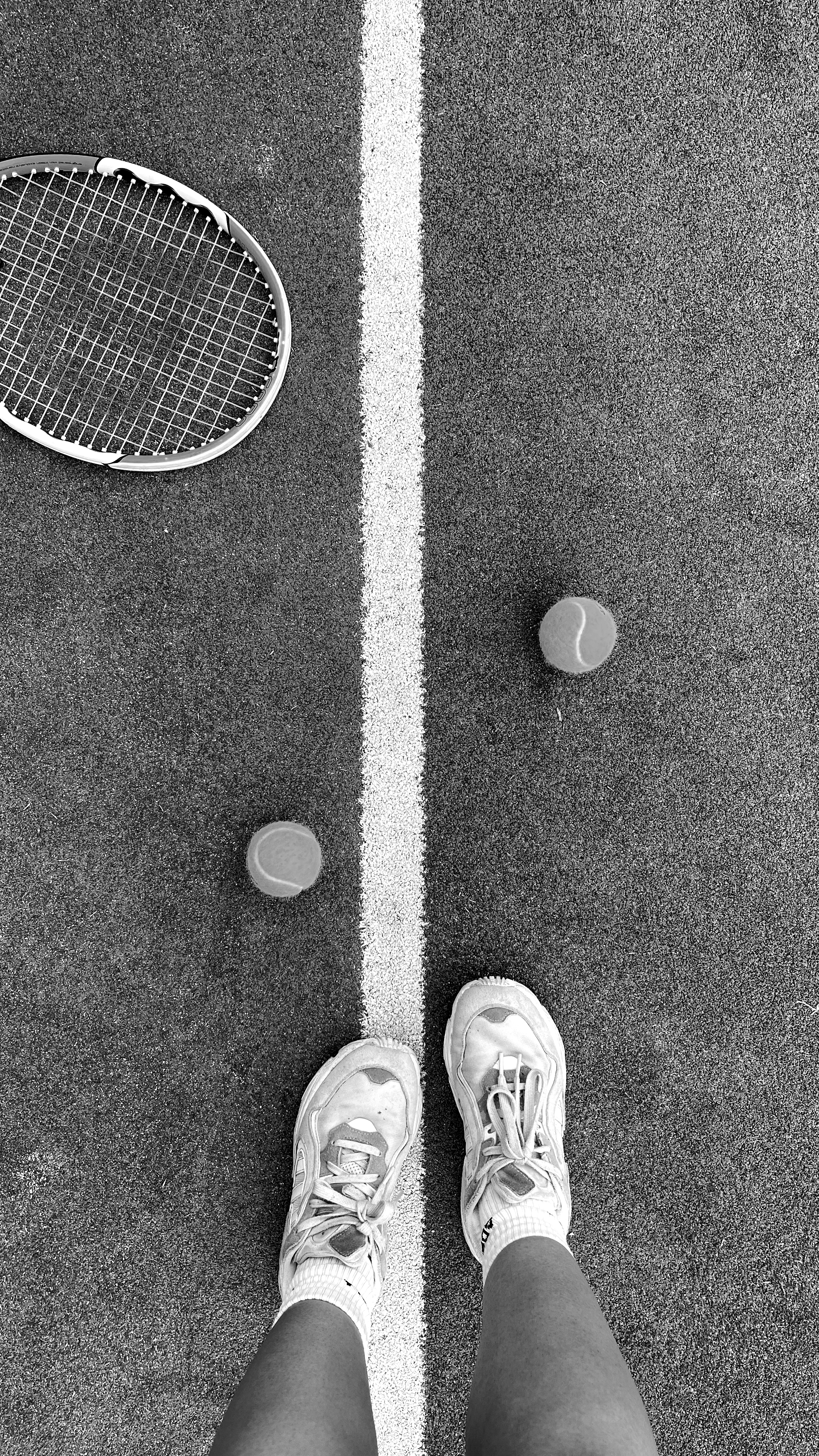 Black and white photo of shoes, ball, and racket on a tennis court.